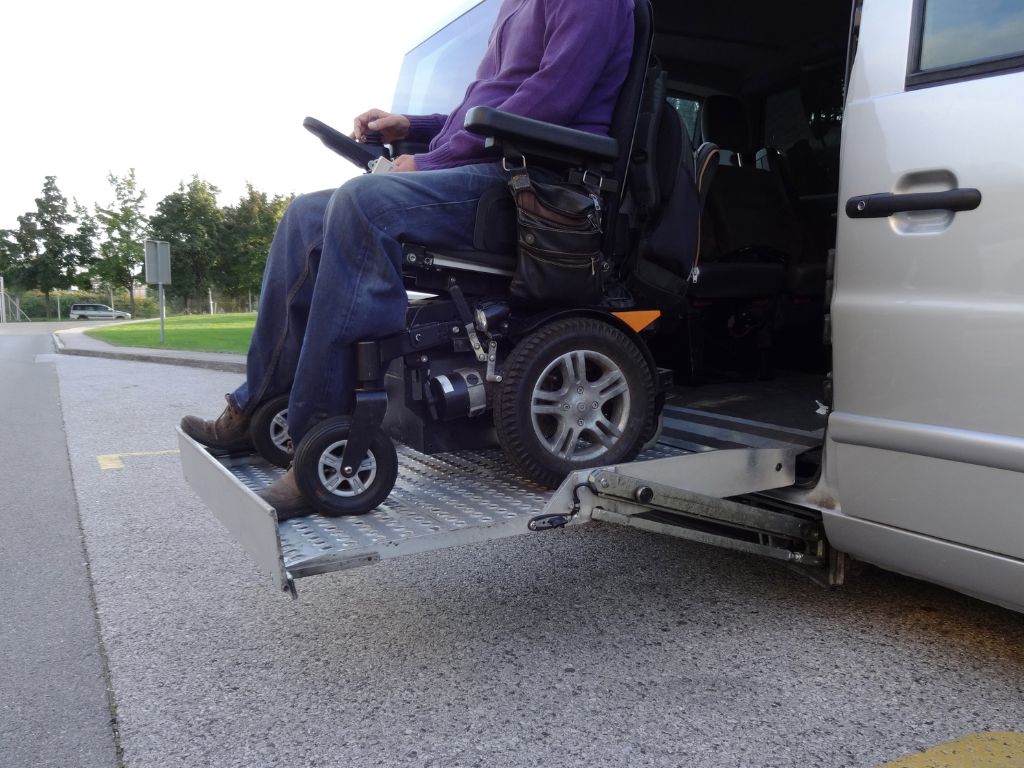 Power wheelchair user with a purple shirt using a metal wheelchair lift to get out of a gray van. 